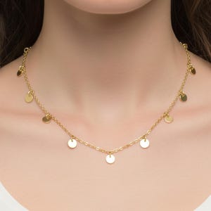 Peut inclure: Collier doré orné de petits disques plats et ronds. Le collier est composé d'une chaîne délicate et est présenté sur le cou d'une personne. C'est un bijou simple et élégant.