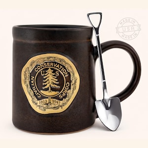 May include: A dark brown ceramic mug with a gold emblem featuring a tree and the text "Civilian Conservation Corps 1933-1942." A silver shovel-shaped spoon rests against the mug. The mug is made in the USA.