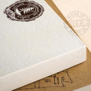 May include: Close-up of two stacked notebooks. The top notebook is off-white with a brown circular emblem that reads "Civilian Conservation Corps" and the years 1933 and 1942. The bottom notebook is brown with a map design.