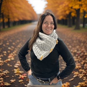 Peut inclure: Une femme portant un col en crochet gris avec des boutons jaunes, un chemisier noir à manches longues et un jean bleu. Elle se tient sur un chemin bordé d'arbres aux feuilles jaunes.