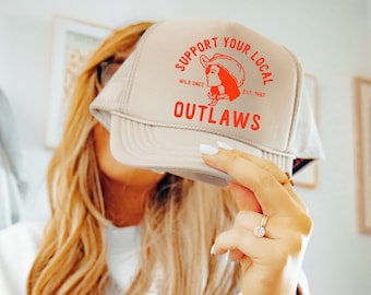 May include: A beige trucker hat with a red and white logo that says "Support Your Local Outlaws" with a drawing of a woman wearing a cowboy hat. The logo also says "Wild Ones Est. 1987".