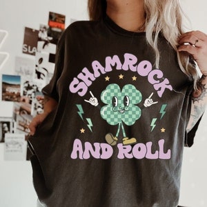 May include: Black t-shirt with a cartoon shamrock wearing yellow shoes and holding up rock and roll hand signs. The text "Shamrock and Roll" is written in a purple font.