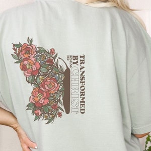 Puede incluir: Una camiseta verde claro con un dise&ntilde;o floral y el texto "TRANSFORMED BY CHRIST" con la referencia b&iacute;blica "ROMANS 12:2" debajo.