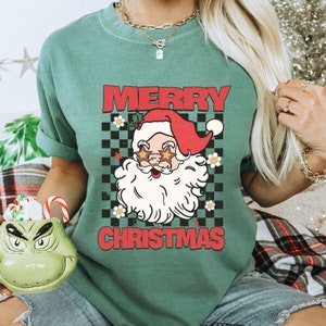 K&ouml;nnte beinhalten: Ein gr&uuml;nes T-Shirt mit einem Vintage-Santa-Claus-Motiv, das eine Sonnenbrille tr&auml;gt. Der Text "Merry Christmas" ist in roten Buchstaben auf dem Shirt gedruckt.