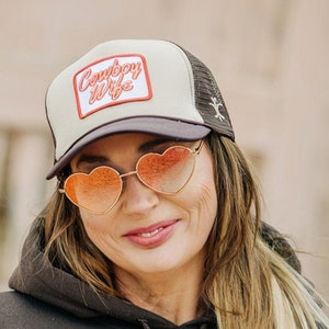 May include: A woman wearing a brown hoodie with a beige and brown trucker hat that says "Cowboy Wife" in red lettering. She is wearing heart-shaped sunglasses with orange lenses.