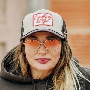 May include: A woman wearing a black hoodie with a white and black trucker hat that says "Country Wife" in red lettering. She is wearing heart-shaped sunglasses.