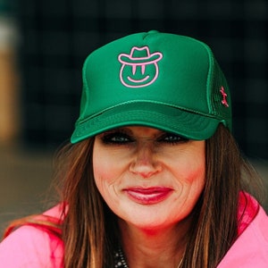May include: A woman wearing a pink shirt and a green trucker hat with a pink smiling face and cowboy hat design.