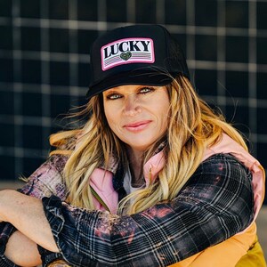 Może przedstawiać: Kobieta w czarnej i różowej czapce trucker z napisem "LUCKY". Ma na sobie koszulę w kratę i wielokolorową kamizelkę.