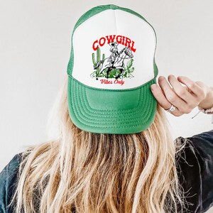 May include: Green and white trucker hat with a cowgirl riding a horse and cactus graphic. The text "COWGIRL Vibes Only" is printed on the hat.
