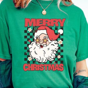 K&ouml;nnte beinhalten: Gr&uuml;nes T-Shirt mit einem Retro-Weihnachtsdesign, das den Weihnachtsmann mit Sonnenbrille und einem karierten Hintergrund zeigt. Der Text "Merry Christmas" ist in roten Buchstaben gedruckt.