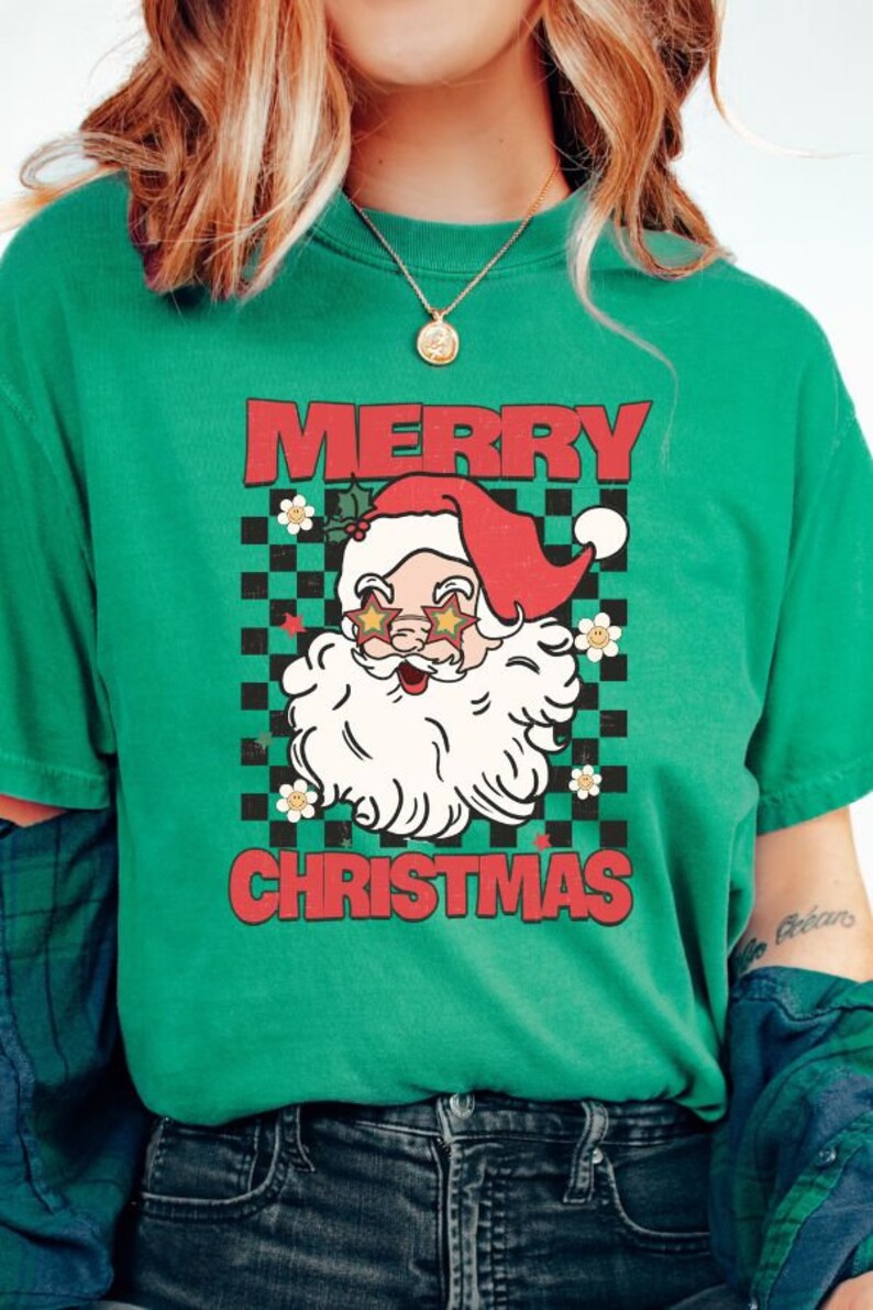 K&ouml;nnte beinhalten: Gr&uuml;nes T-Shirt mit einem Retro-Weihnachtsdesign, das den Weihnachtsmann mit Sonnenbrille und einem karierten Hintergrund zeigt. Der Text "Merry Christmas" ist in roten Buchstaben gedruckt.