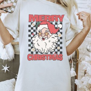 K&ouml;nnte beinhalten: Wei&szlig;es T-Shirt mit einem rot-wei&szlig; karierten Hintergrund, einem Cartoon-Weihnachtsmann mit Sonnenbrille und dem Text "Frohe Weihnachten".