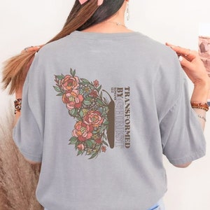 Puede incluir: Una camiseta gris con un dise&ntilde;o de mariposa floral y el texto "TRANSFORMED BY CHRIST" con "ROMANS 12:2" debajo.