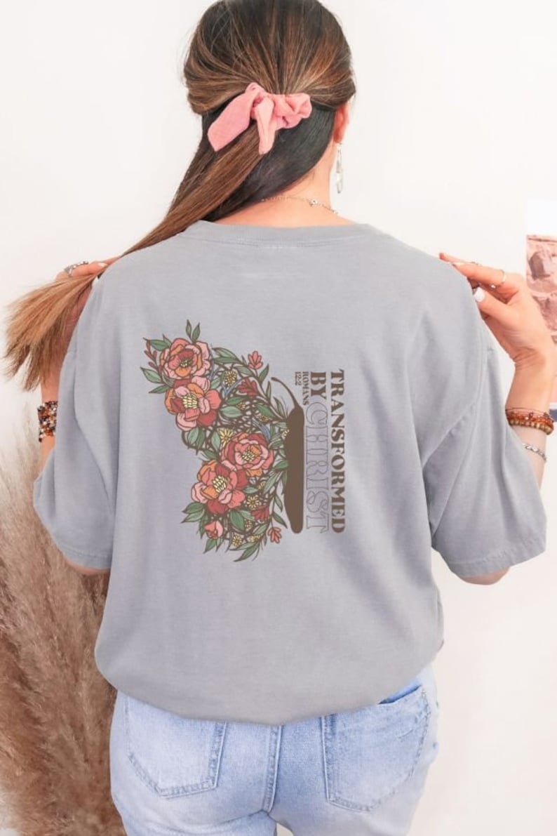 Puede incluir: Una camiseta gris con un dise&ntilde;o de mariposa floral y el texto "TRANSFORMED BY CHRIST" con "ROMANS 12:2" debajo.