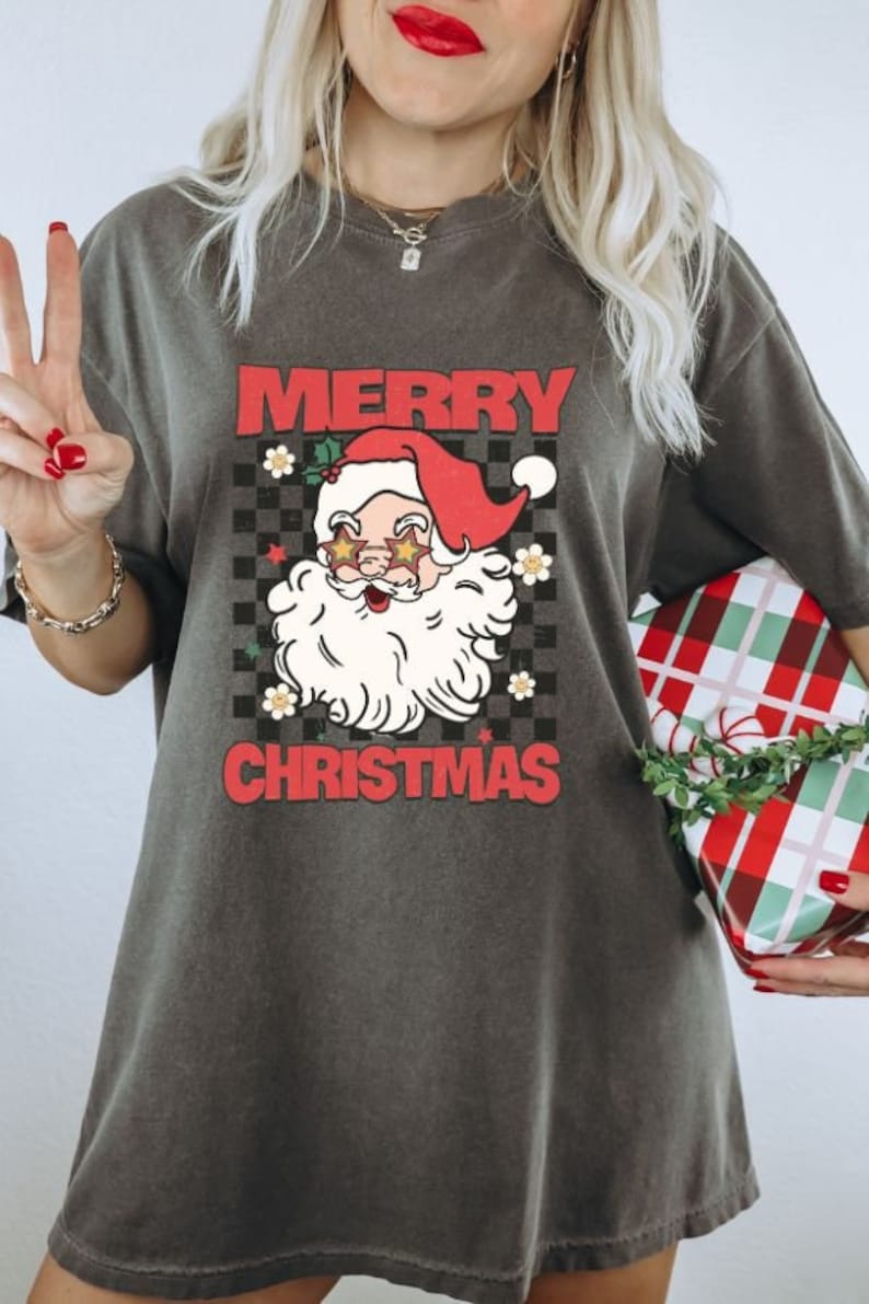 K&ouml;nnte beinhalten: Ein dunkelgraues T-Shirt mit einer Vintage-Illustration von Weihnachtsmann mit Sonnenbrille und roter M&uuml;tze. Der Text "Merry Christmas" ist in roten Buchstaben auf dem Shirt gedruckt.