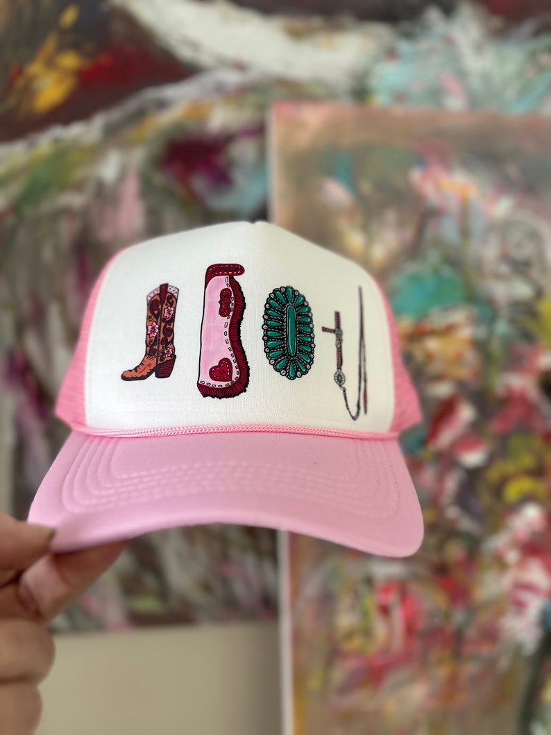 May include: Pink trucker hat with a white front panel featuring a graphic of a cowboy boot, a riding boot, a turquoise and silver concho, and a riding crop.