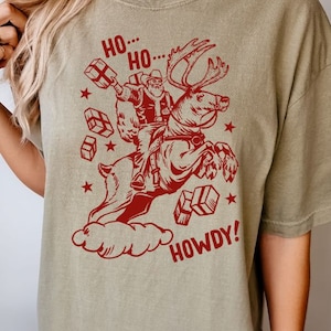 May include: A green t-shirt with a red graphic of Santa Claus riding a reindeer and holding presents. The text "Ho... Ho... Howdy!" is printed on the shirt.