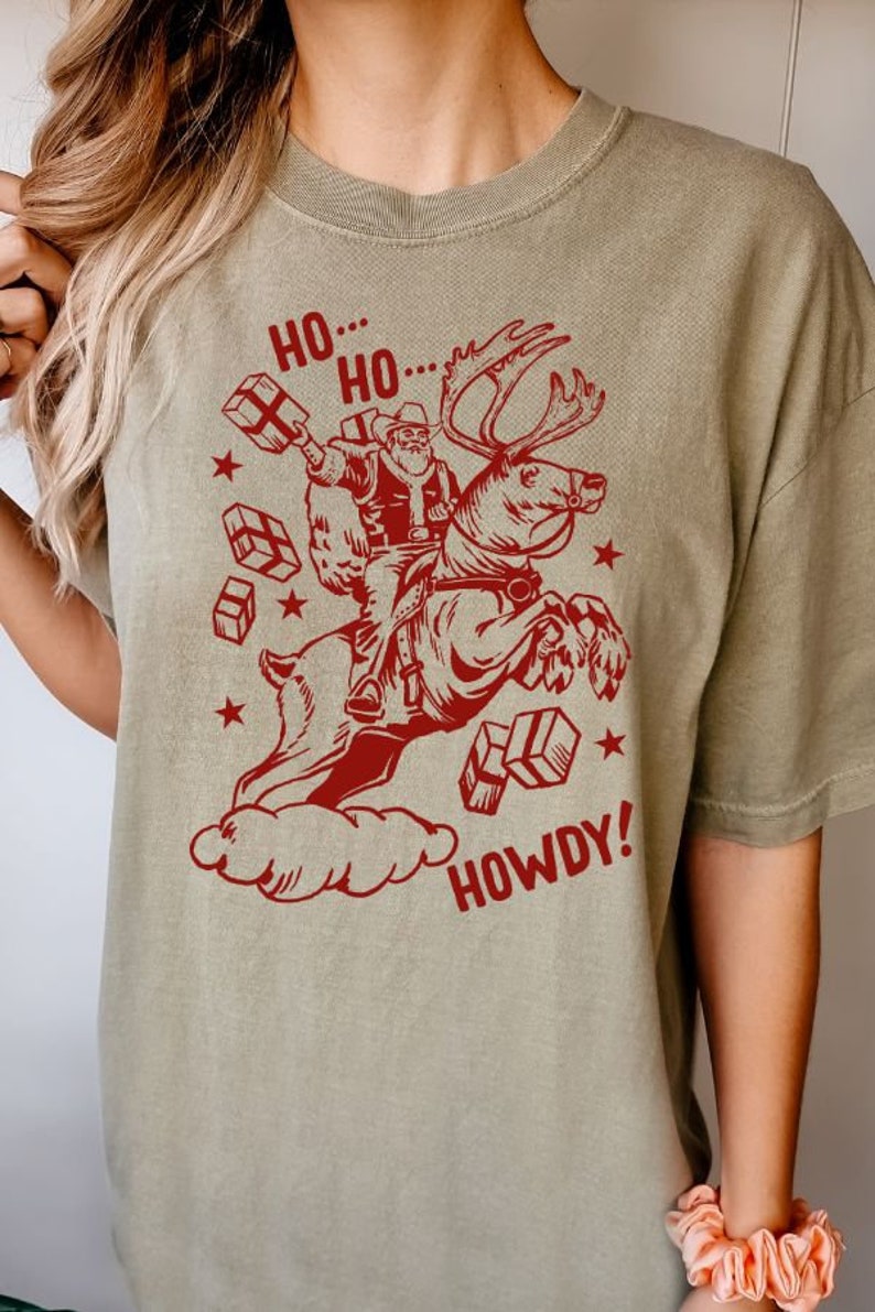 May include: A green t-shirt with a red graphic of Santa Claus riding a reindeer and holding presents. The text "Ho... Ho... Howdy!" is printed on the shirt.