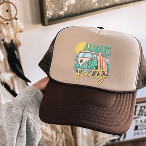 May include: A brown and beige trucker hat with a white van and surfboards graphic. The text "Always Salty" is printed on the hat.