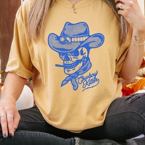 May include: A mustard yellow t-shirt featuring a blue graphic of a skull wearing a cowboy hat and bandana. The text "Cowboy Killer" is written in blue cursive below the skull.