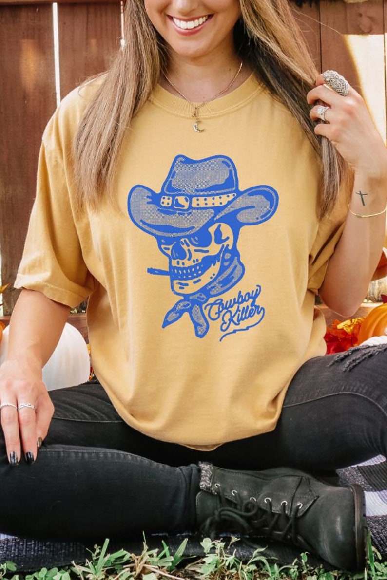 May include: A mustard yellow t-shirt featuring a blue graphic of a skull wearing a cowboy hat and bandana. The text "Cowboy Killer" is written in blue cursive below the skull.