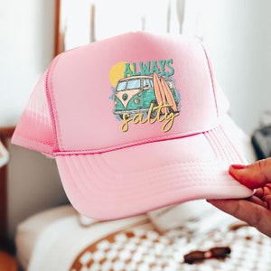 May include: Pink trucker hat with a white mesh back. The front of the hat features a graphic of a vintage van with surfboards and the text "Always Salty".