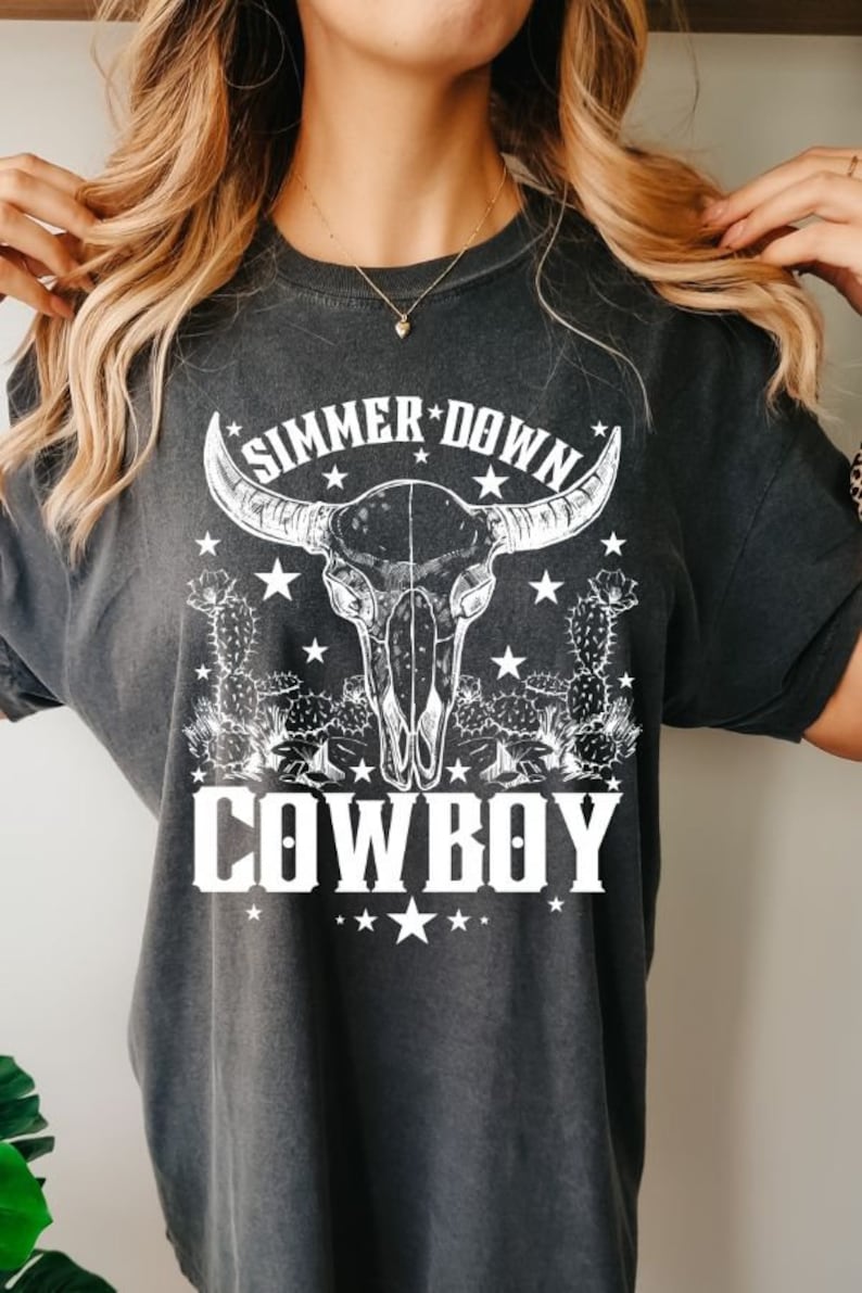 May include: A dark gray t-shirt with a white graphic design of a bull skull with cactus plants and stars. The text "Simmer Down Cowboy" is printed below the skull.