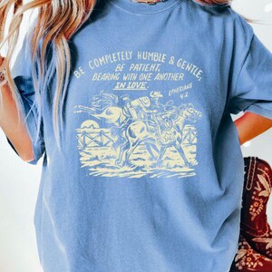 K&ouml;nnte beinhalten: Ein hellblaues T-Shirt mit einer Vintage-Grafik eines Cowboys, der ein Pferd reitet. Die Grafik enth&auml;lt den Text "Be completely humble & gentle. Be patient. Bearing with one another in love. Ephesians 4:2."