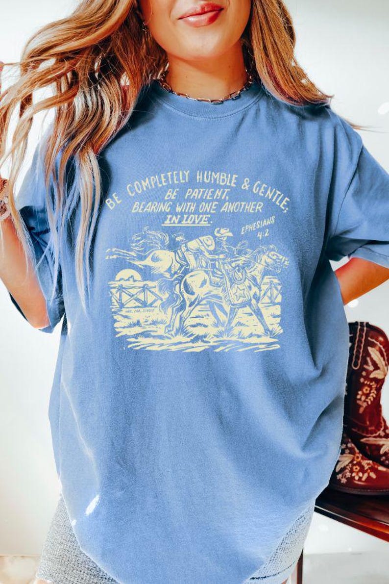 K&ouml;nnte beinhalten: Ein hellblaues T-Shirt mit einer Vintage-Grafik eines Cowboys, der ein Pferd reitet. Die Grafik enth&auml;lt den Text "Be completely humble & gentle. Be patient. Bearing with one another in love. Ephesians 4:2."