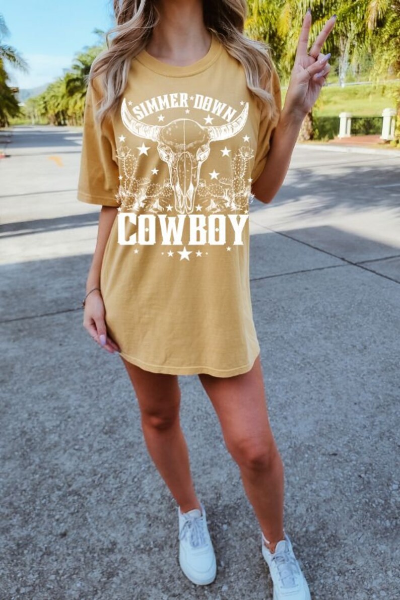 May include: A yellow t-shirt with a white graphic of a skull with horns, cacti, and stars. The text "Summer Down Cowboy" is printed on the shirt.