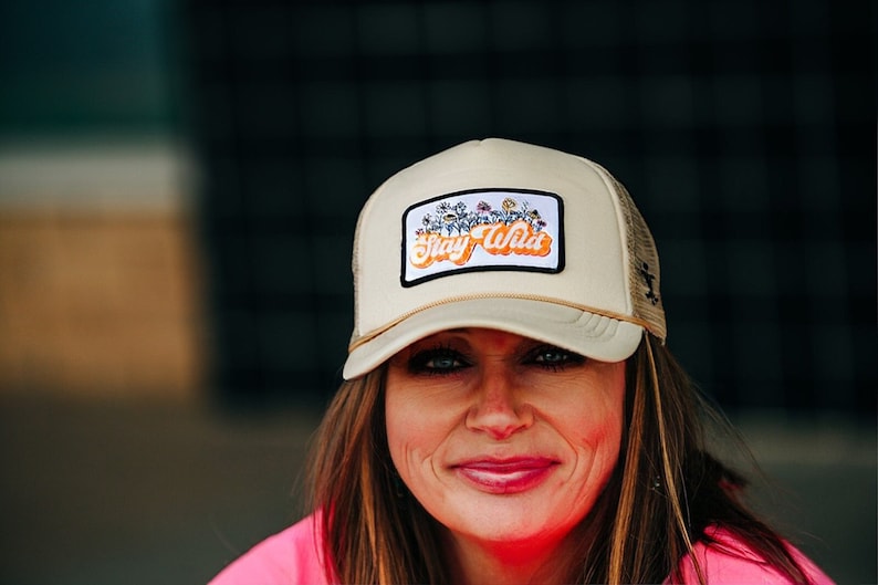 May include: A beige trucker hat with a patch that says "Stay Wild" in orange letters with a floral design. The patch has a white border.