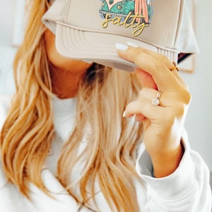 May include: A beige trucker hat with a white and green graphic of a vintage van with surfboards and the text "Always Salty".
