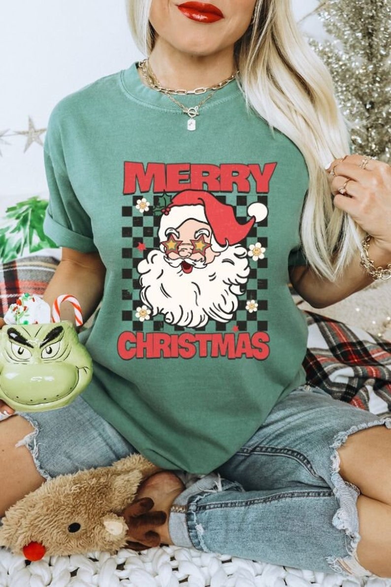 K&ouml;nnte beinhalten: Ein gr&uuml;nes T-Shirt mit einem Vintage-Santa-Claus-Motiv, das eine Sonnenbrille tr&auml;gt. Der Text "Merry Christmas" ist in roten Buchstaben auf dem Shirt gedruckt.