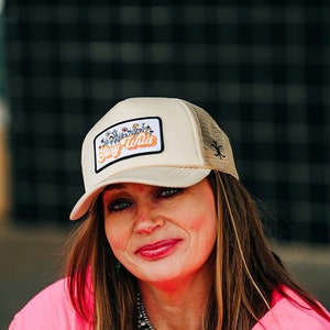 May include: A woman wearing a pink shirt and a beige trucker hat with a floral patch that says "Stay Wild".