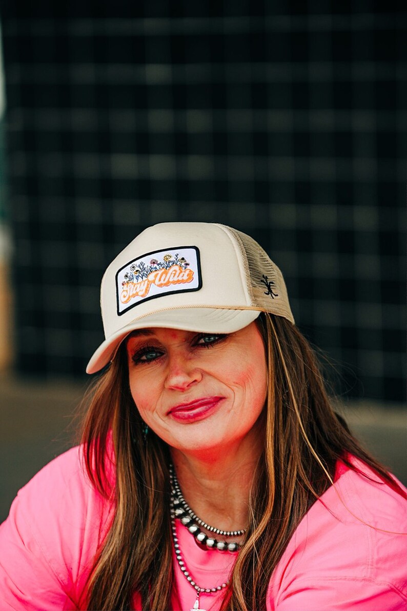 May include: A woman wearing a pink shirt and a beige trucker hat with a floral patch that says "Stay Wild".
