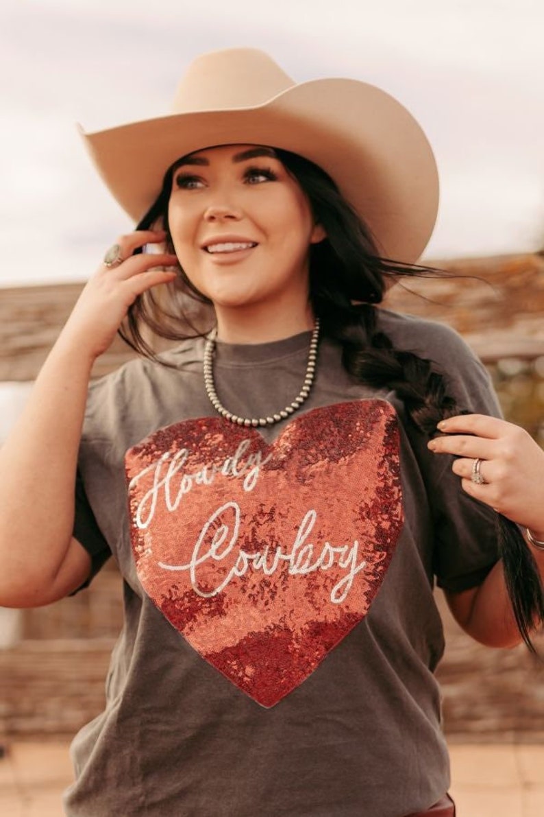 May include: A gray t-shirt with a sequined heart that says "Howdy Cowgirl" in white lettering. The model is wearing a brown cowboy hat and a silver necklace.