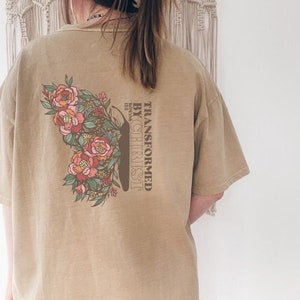 Puede incluir: Una camiseta de color marr&oacute;n claro con un dise&ntilde;o floral y el texto "TRANSFORMED BY CHRIST" con la referencia b&iacute;blica "ROMANS 12:2".