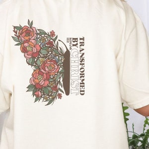 Puede incluir: Una camiseta de color crema con un dise&ntilde;o de mariposa floral y el texto "TRANSFORMED BY CHRIST" con la referencia b&iacute;blica "ROMANS 12:2".
