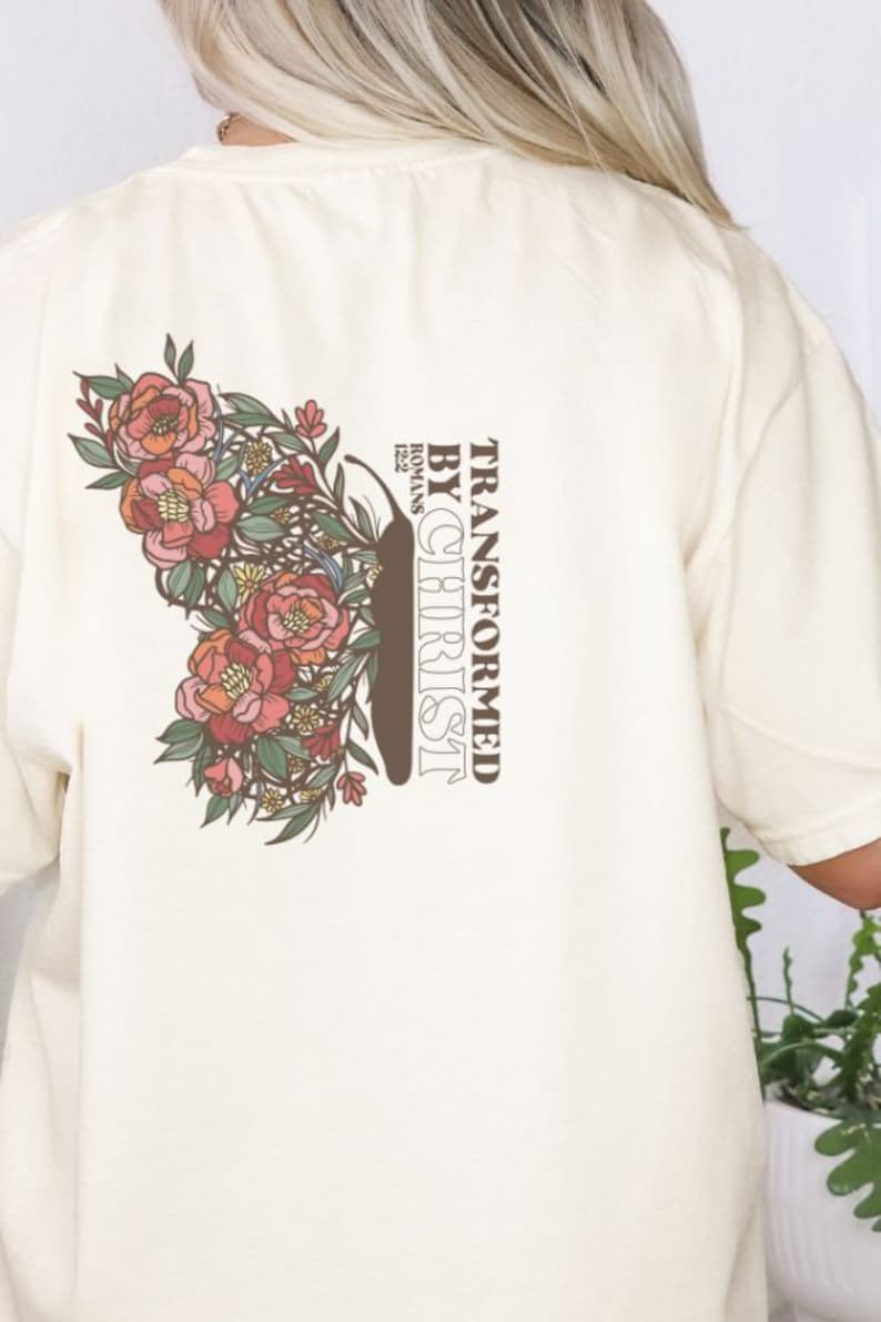 Puede incluir: Una camiseta de color crema con un dise&ntilde;o de mariposa floral y el texto "TRANSFORMED BY CHRIST" con la referencia b&iacute;blica "ROMANS 12:2".