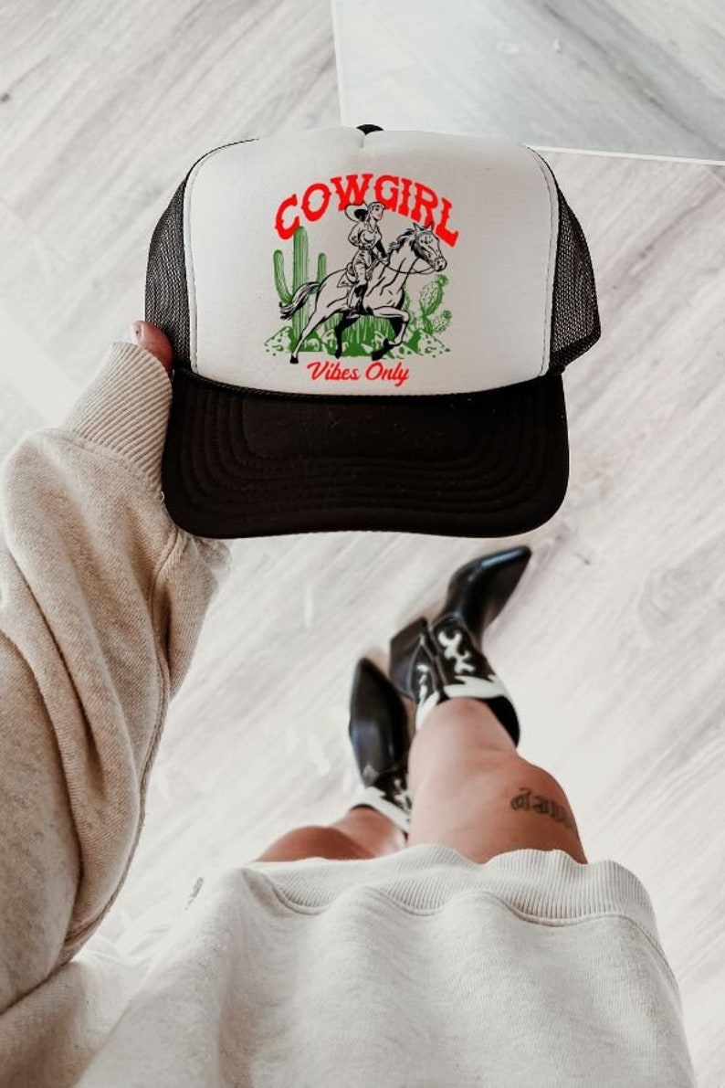 May include: White trucker hat with black mesh back. The hat has a black and white illustration of a cowgirl riding a horse with cacti and the text "COWGIRL Vibes Only".