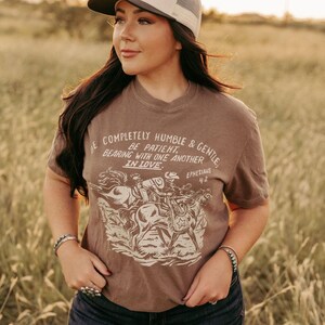 K&ouml;nnte beinhalten: Ein braunes T-Shirt mit einer Grafik von Cowboys, die auf Pferden reiten. Der Text auf dem Shirt lautet "Be completely humble & gentle, Be patient, Bearing with one another in love, Ephesians 4:2".
