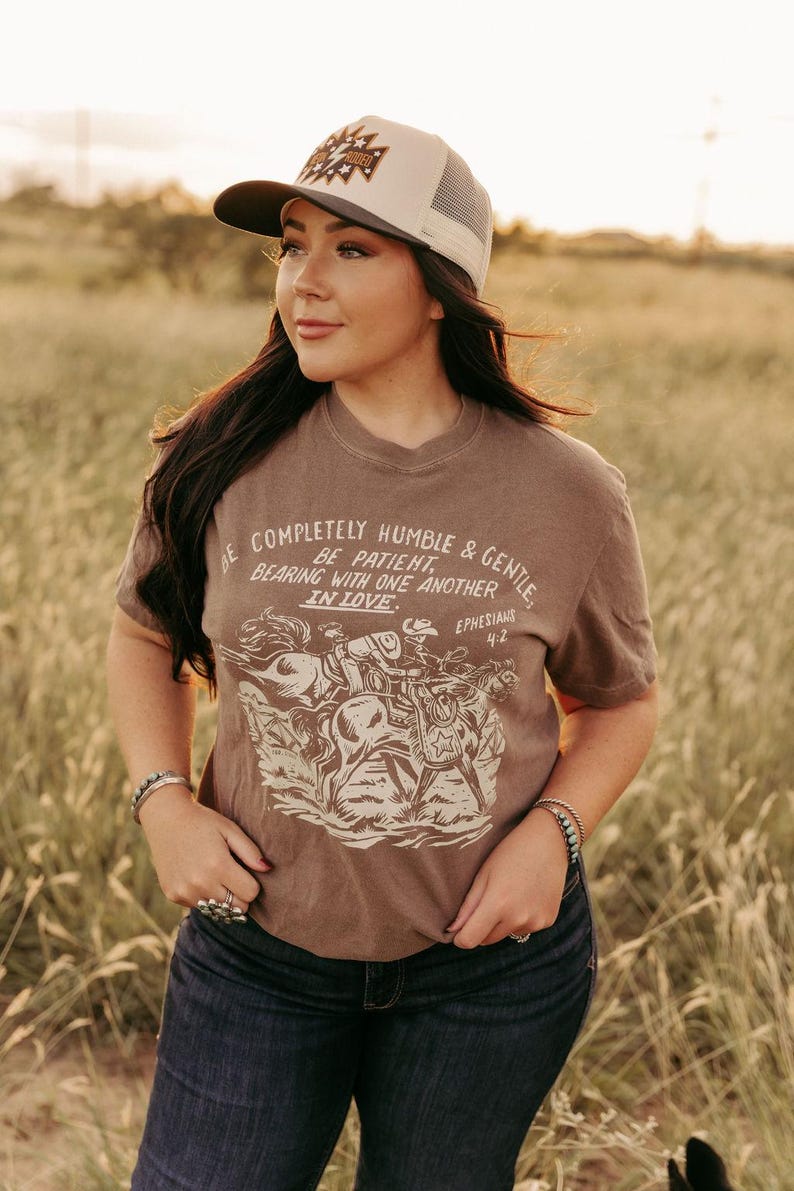 K&ouml;nnte beinhalten: Ein braunes T-Shirt mit einer Grafik von Cowboys, die auf Pferden reiten. Der Text auf dem Shirt lautet "Be completely humble & gentle, Be patient, Bearing with one another in love, Ephesians 4:2".