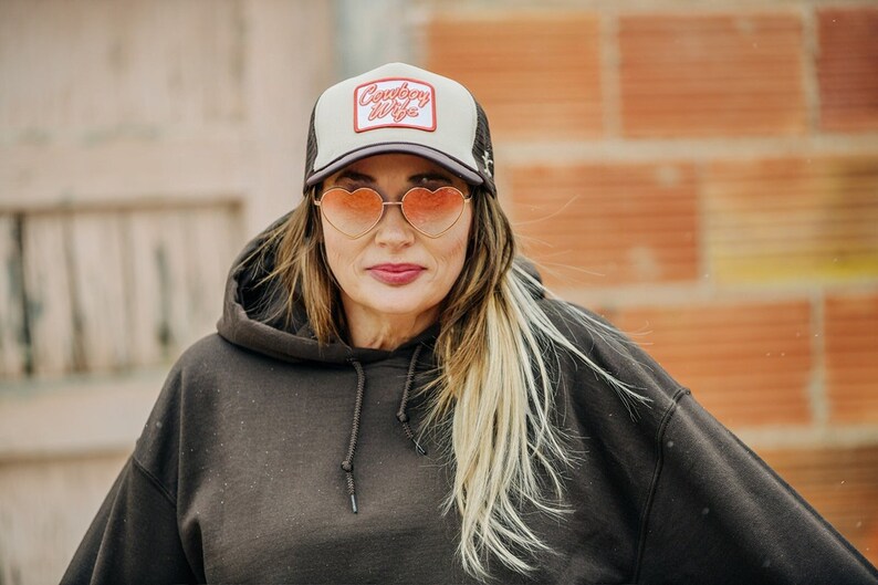 May include: A woman wearing a black hoodie with a white and black trucker hat that says "Country Wife" in red lettering. She is wearing heart-shaped sunglasses.