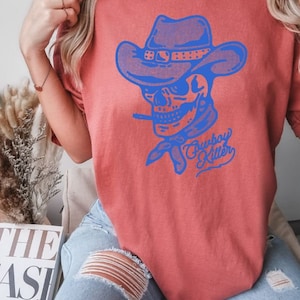 May include: A red t-shirt featuring a blue graphic of a skull wearing a cowboy hat and bandana. The text "Cowboy Killer" is printed beneath the skull.