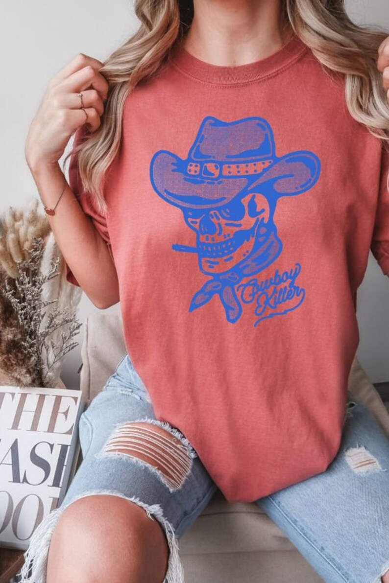 May include: A red t-shirt featuring a blue graphic of a skull wearing a cowboy hat and bandana. The text "Cowboy Killer" is printed beneath the skull.