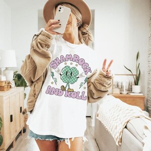 May include: White t-shirt with a green shamrock cartoon character wearing a checkered hat and holding up peace signs. The text "Shamrock and Roll" is written in purple letters around the shamrock.