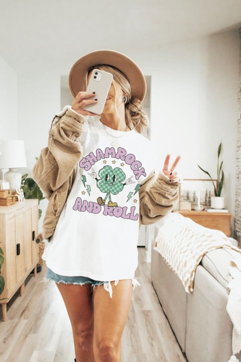 May include: White t-shirt with a green shamrock cartoon character wearing a checkered hat and holding up peace signs. The text "Shamrock and Roll" is written in purple letters around the shamrock.