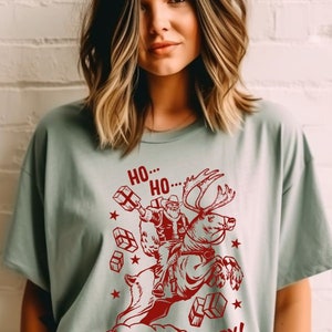 May include: A light green t-shirt with a red graphic of Santa Claus riding a reindeer and holding presents. The text "Ho... Ho... Howdy!" is printed on the shirt.