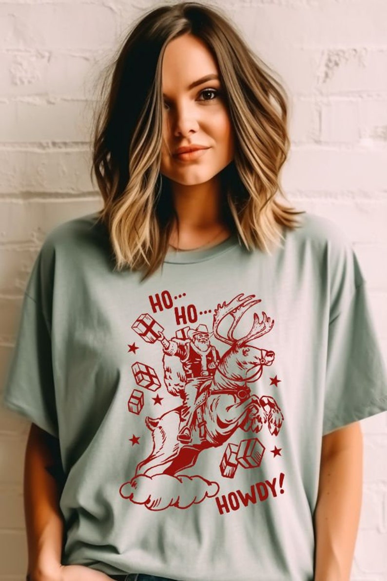 May include: A light green t-shirt with a red graphic of Santa Claus riding a reindeer and holding presents. The text "Ho... Ho... Howdy!" is printed on the shirt.