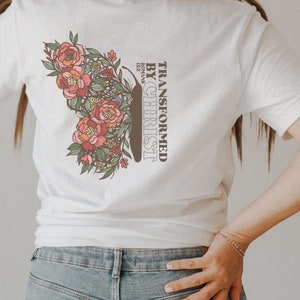 Puede incluir: Camiseta blanca con un dise&ntilde;o floral y el texto "Transformado por Cristo" con la referencia b&iacute;blica "Romanos 12:2" debajo.